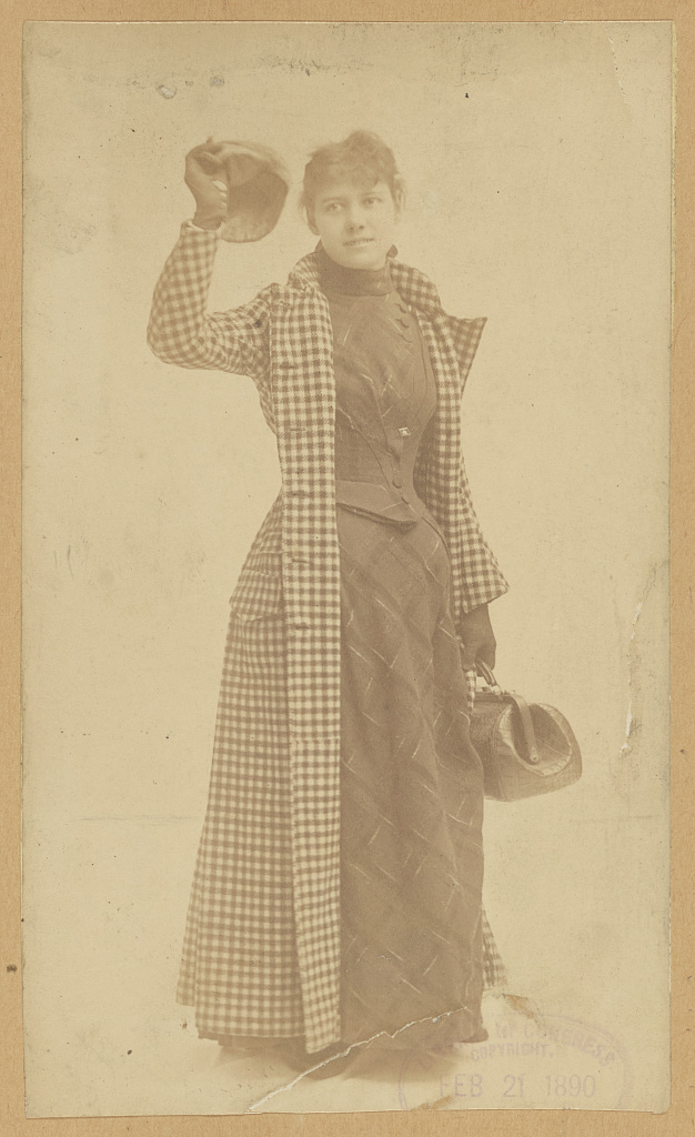 Nellie Bly standing with a hat and bag.